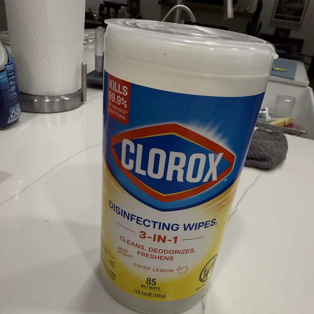 A rough phone snapshot of a Clorox disinfecting wipes canister on a cluttered kitchen counter, shot under flat overhead light next to a paper towel roll and a dish rag